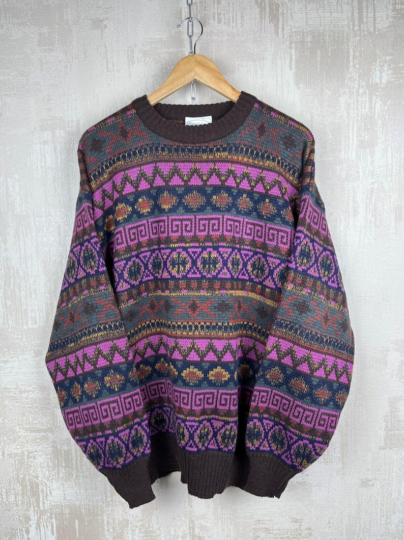 K&ouml;nnte beinhalten: Ein Vintage-Pullover mit Rundhalsausschnitt und geometrischem Muster in Braun-, Lila-, Blau- und Gelbt&ouml;nen. Der Pullover h&auml;ngt an einem Holzb&uuml;gel. Das Design umfasst Zickzack-, Quadrat- und Diamantformen.
