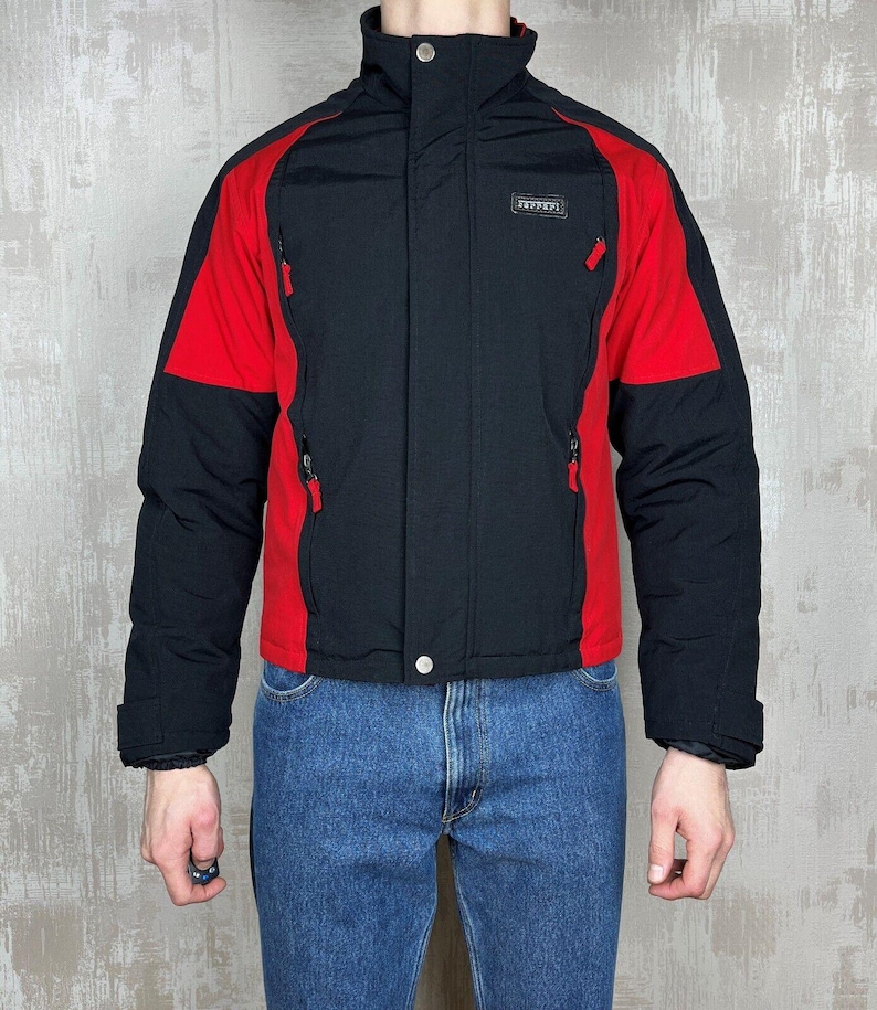 May include: Black and red Ferrari jacket with a high collar and zippered pockets. The jacket has a black body with red shoulder and sleeve accents. The Ferrari logo is visible on the chest.