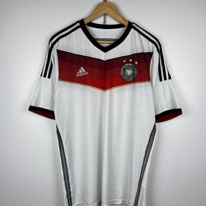 May include: White soccer jersey with black and red stripes, Adidas logo, and the German national team crest. The jersey has a V-neck and short sleeves.