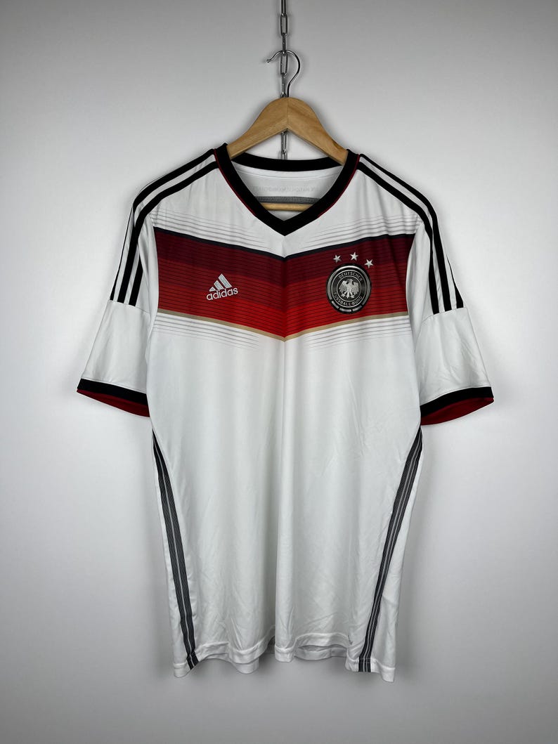 May include: White soccer jersey with black and red stripes, Adidas logo, and the German national team crest. The jersey has a V-neck and short sleeves.