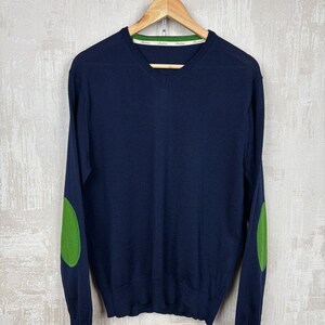 Könnte beinhalten: Ein dunkelblauer V-Ausschnitt-Pullover mit langen Ärmeln und leuchtend grünen Ellbogen-Patches. Der innere Halsausschnitt ist mit einem weiß-grünen Besatz versehen. Der Pullover hängt auf einem Holzkleiderbügel vor einem hellgrauen Hintergrund.