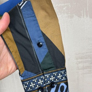 May include: Close-up of a patterned shirt sleeve. The sleeve features a geometric design in shades of blue, brown, black, and green. The cuff has a decorative pattern with black buttons. The shirt is made of a smooth fabric.