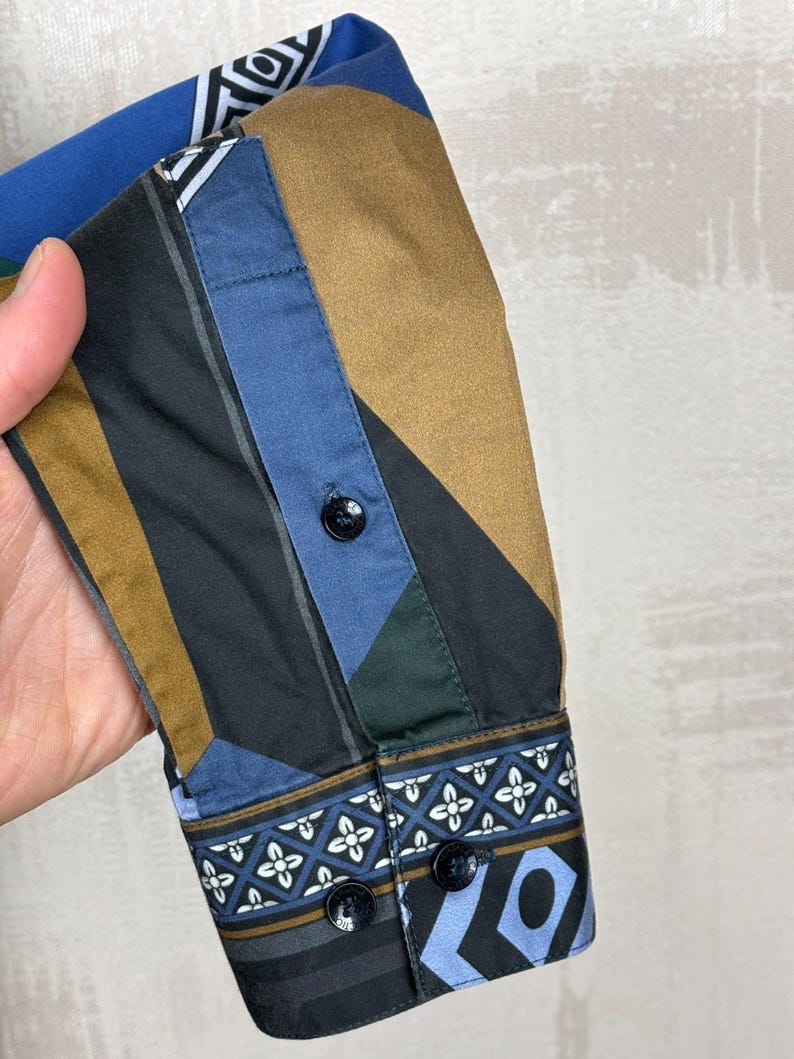 May include: Close-up of a patterned shirt sleeve. The sleeve features a geometric design in shades of blue, brown, black, and green. The cuff has a decorative pattern with black buttons. The shirt is made of a smooth fabric.