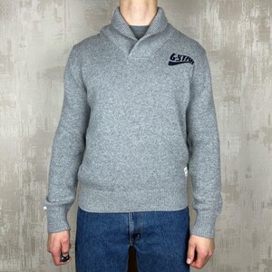 Könnte beinhalten: Ein grauer Pullover mit Schalkragen und langen Ärmeln. Der Pullover hat den marineblauen Text "G-Star" auf der oberen Brust. Das Model trägt eine blaue Jeans.