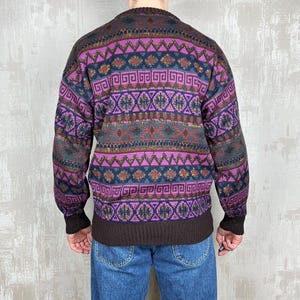 K&ouml;nnte beinhalten: Ein gemusterter Pullover in Lila-, Braun- und Marineblaut&ouml;nen. Der Pullover hat geometrische Muster und einen gerippten Saum und B&uuml;ndchen. Die Person tr&auml;gt eine blaue Jeans.