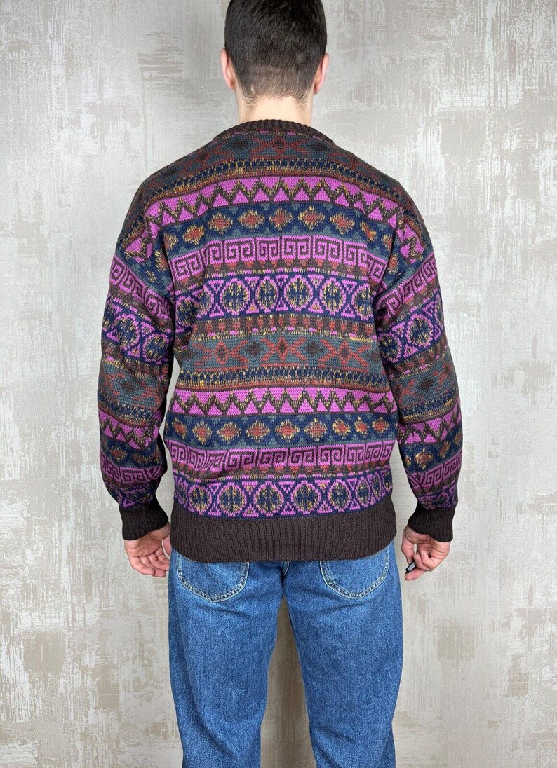 K&ouml;nnte beinhalten: Ein gemusterter Pullover in Lila-, Braun- und Marineblaut&ouml;nen. Der Pullover hat geometrische Muster und einen gerippten Saum und B&uuml;ndchen. Die Person tr&auml;gt eine blaue Jeans.