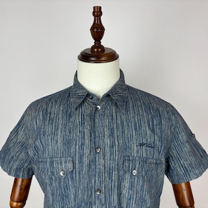 Puede incluir: Una camisa de manga corta con botones en un patrón de textura vertical azul y gris. La camisa presenta un cuello estándar, una tapeta frontal con botones y dos bolsillos en el pecho con solapas abotonadas. Un pequeño logotipo azul oscuro está bordado en el pecho.