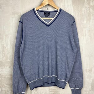 Puede incluir: Un suéter de cuello en V a rayas azules y blancas con mangas largas, colgado de una percha de madera. El suéter tiene ribetes blancos a lo largo del escote, los puños y el dobladillo. La marca "HACKETT" es visible en una etiqueta en el escote.