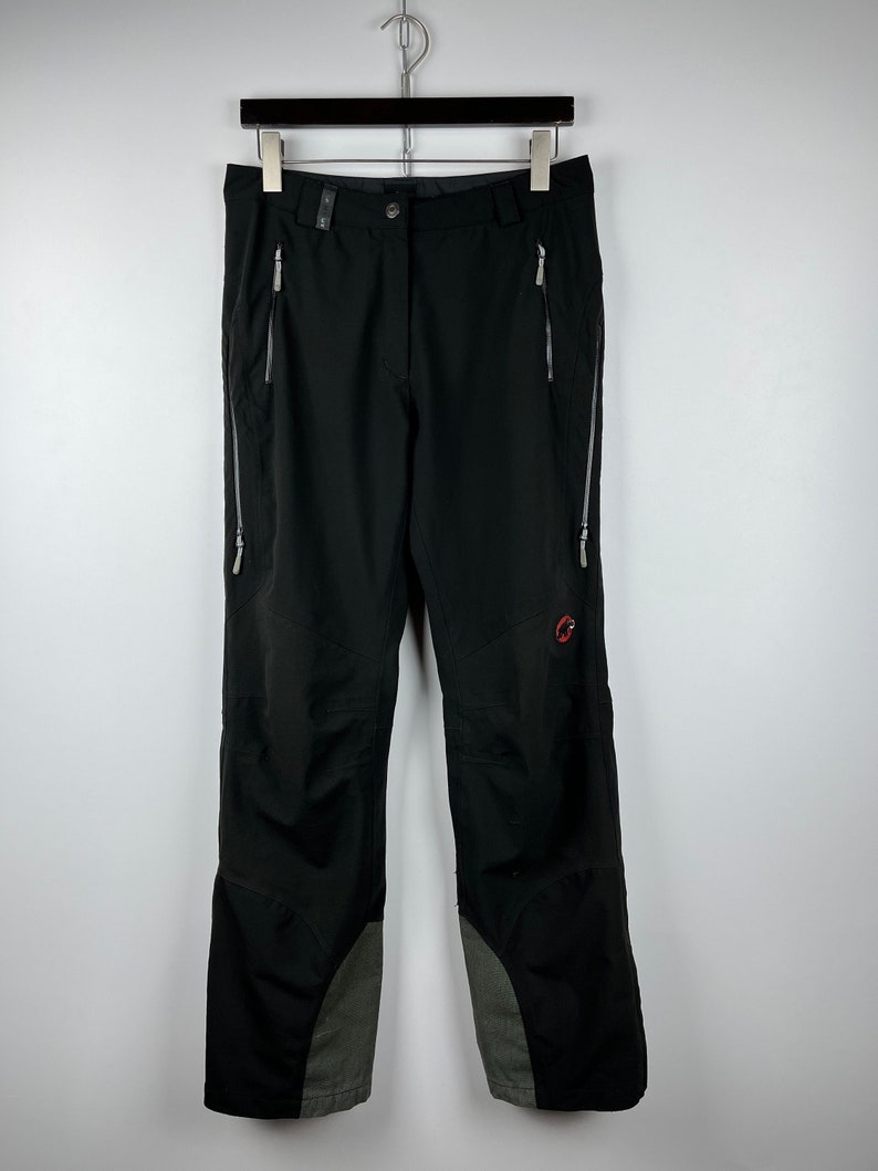 May include: Black hiking pants with zippered pockets and a red logo on the right leg. The pants have a gray panel on the lower legs.