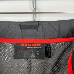 May include: A black and red Mammut brand clothing size tag with the text "Swiss Technology" and size information in European, UK, USA, and Asian sizing. The tag reads: EU/D 40, UK 14, F 42, USA 10, 44, ASIA L.