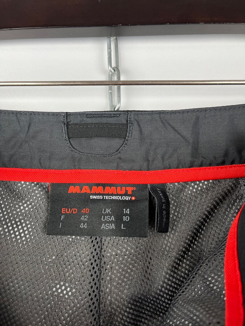 May include: A black and red Mammut brand clothing size tag with the text "Swiss Technology" and size information in European, UK, USA, and Asian sizing. The tag reads: EU/D 40, UK 14, F 42, USA 10, 44, ASIA L.