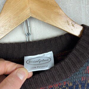K&ouml;nnte beinhalten: Ein Vintage-Pullover mit braunem Rippstrick-Ausschnitt und -B&uuml;ndchen. Der Pullover hat ein farbenfrohes geometrisches Muster in Blau-, Gelb-, Rot- und Rosat&ouml;nen. Das Etikett lautet "Enrico Gorlani" und "100% Polyacryl". Der Pullover h&auml;ngt an einem Holzkleiderb&uuml;gel.