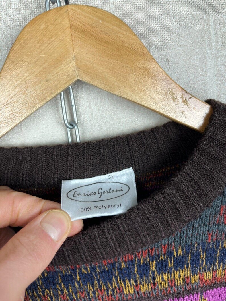 K&ouml;nnte beinhalten: Ein Vintage-Pullover mit braunem Rippstrick-Ausschnitt und -B&uuml;ndchen. Der Pullover hat ein farbenfrohes geometrisches Muster in Blau-, Gelb-, Rot- und Rosat&ouml;nen. Das Etikett lautet "Enrico Gorlani" und "100% Polyacryl". Der Pullover h&auml;ngt an einem Holzkleiderb&uuml;gel.