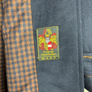 May include: A navy blue coat with a brown and black plaid lining. The coat has a label that reads "Original Alpenloden" with a crest and the text "Original Alpenloden" in white.