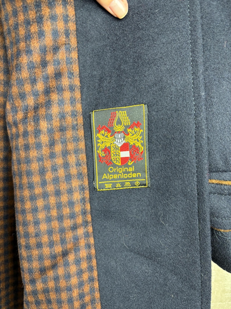 May include: A navy blue coat with a brown and black plaid lining. The coat has a label that reads "Original Alpenloden" with a crest and the text "Original Alpenloden" in white.