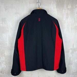 May include: Black jacket with red side panels. The jacket has a stand-up collar and a small red design on the upper back. The jacket is hanging from a wooden hanger.