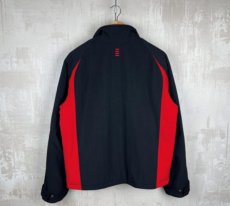 May include: Black jacket with red side panels. The jacket has a stand-up collar and a small red design on the upper back. The jacket is hanging from a wooden hanger.