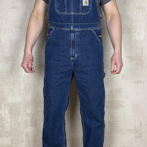 Może przedstawiać: Niebieskie dżinsowe ogrodniczki z regulowanymi ramiączkami i wieloma kieszeniami. Mała naszywka z logo na kieszeni na piersi. Osoba ma na sobie szary t-shirt i szare trampki.