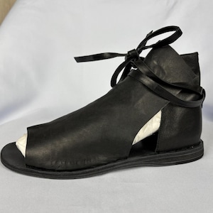 May include: Black leather sandal with an open toe design. The sandal features a strap that wraps around the ankle and ties in a bow. The shoe has a flat sole and a cutout design on the sides.