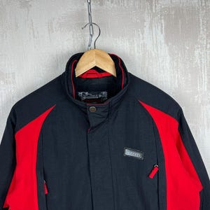 May include: Black and red jacket with a high collar, hanging from a wooden hanger. The jacket has red panels on the shoulders and sides, with a small rectangular patch on the chest. The jacket has two zippered pockets.