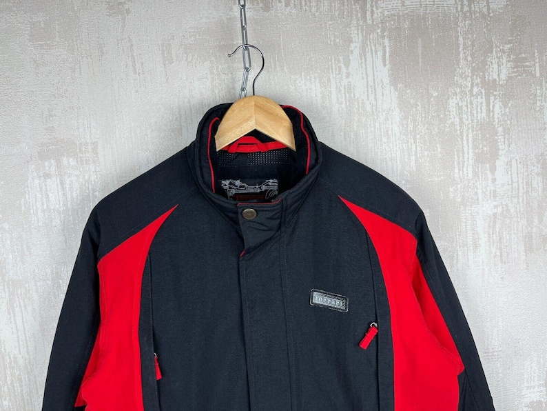 May include: Black and red jacket with a high collar, hanging from a wooden hanger. The jacket has red panels on the shoulders and sides, with a small rectangular patch on the chest. The jacket has two zippered pockets.