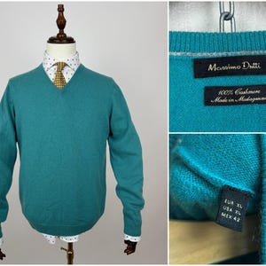 May include: Teal V-neck sweater on a mannequin. The sweater is made of 100% cashmere and has long sleeves. The label reads "Massimo Dutti" and "Made in Madagascar". The mannequin is wearing a white shirt with a polka dot pattern and a yellow tie.
