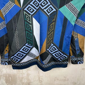 May include: A long-sleeved shirt with a geometric pattern in shades of blue, green, black, and brown. The design features squares and diamond shapes. The cuffs and collar have a contrasting border.