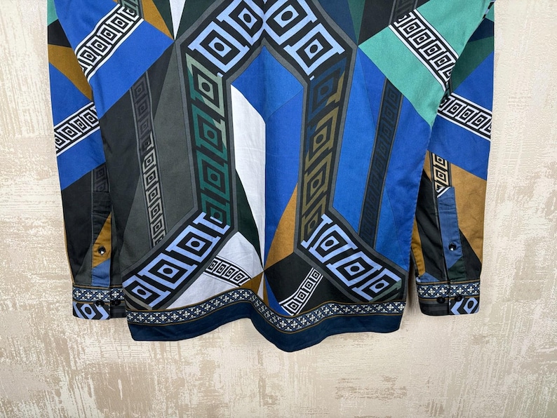 May include: A long-sleeved shirt with a geometric pattern in shades of blue, green, black, and brown. The design features squares and diamond shapes. The cuffs and collar have a contrasting border.