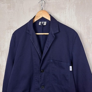 Puede incluir: Blazer azul marino con solapas de muesca, tres botones negros y un bolsillo en el pecho. La chaqueta está colgada de una percha de madera sobre un fondo neutro. Se ve una pequeña etiqueta blanca.