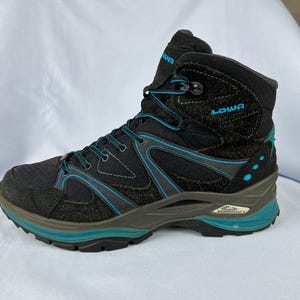 Puede incluir: Botas de senderismo negras con detalles azules y la palabra "LOWA" en el lateral. Las botas tienen una suela exterior robusta y un cuello de tobillo acolchado.