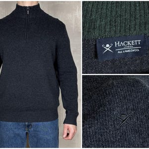 Puede incluir: Jersey gris oscuro con cremallera y cuello de canalé. El jersey tiene un cuello alto y una cremallera que llega hasta el pecho. La etiqueta Hackett London es visible. Hecho de lana de cordero.