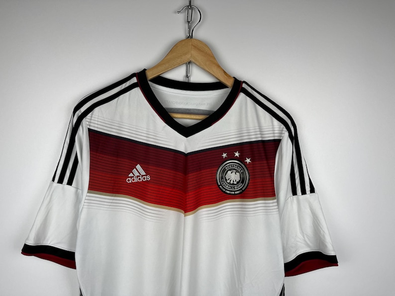 May include: White soccer jersey with black and red stripes, the Adidas logo, and the German national team crest. The jersey has a V-neck collar and short sleeves.