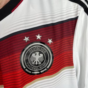 May include: White soccer jersey with red and black stripes. The jersey features the German national team crest, which includes a black eagle with a silver outline and the text "DEUTSCHER FUSSBALL-BUND".