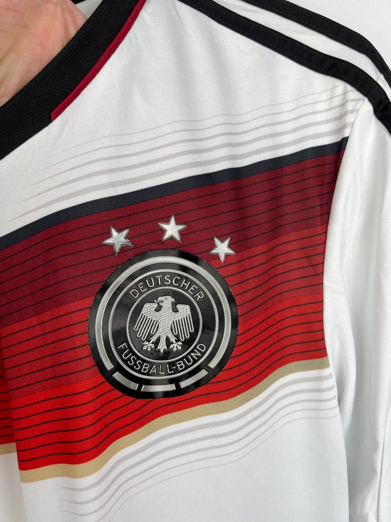 May include: White soccer jersey with red and black stripes. The jersey features the German national team crest, which includes a black eagle with a silver outline and the text "DEUTSCHER FUSSBALL-BUND".