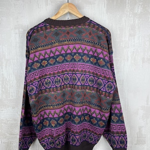 K&ouml;nnte beinhalten: Ein Vintage-Pullover mit farbenfrohem geometrischem Muster. Der Pullover hat einen braunen Ausschnitt, B&uuml;ndchen und Saum. Der K&ouml;rper des Pullovers hat ein Muster aus lila, blauen, gr&uuml;nen und goldenen geometrischen Formen. Der Pullover h&auml;ngt an einem Holzb&uuml;gel.