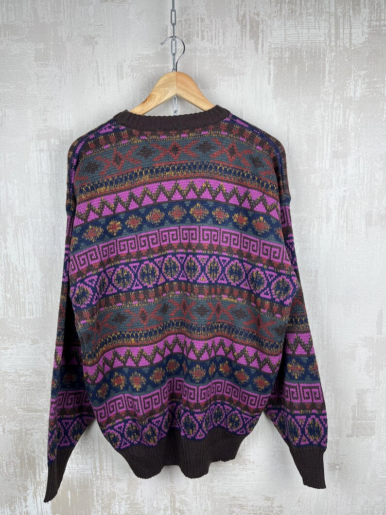 K&ouml;nnte beinhalten: Ein Vintage-Pullover mit farbenfrohem geometrischem Muster. Der Pullover hat einen braunen Ausschnitt, B&uuml;ndchen und Saum. Der K&ouml;rper des Pullovers hat ein Muster aus lila, blauen, gr&uuml;nen und goldenen geometrischen Formen. Der Pullover h&auml;ngt an einem Holzb&uuml;gel.
