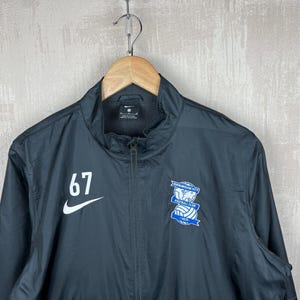 Könnte beinhalten: Schwarze Nike Jacke mit durchgehendem Reißverschluss, mit der weißen Nummer "67" auf der linken Brust. Ein weißer Nike Swoosh befindet sich unter der Nummer. Das Logo des Birmingham City Football Club befindet sich auf der rechten Brust.
