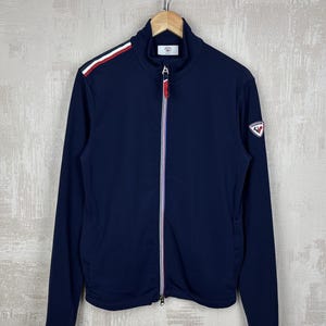 May include: Navy blue zip-up jacket with a high collar. The jacket features a white, red, and green stripe detail on the shoulder and a logo patch on the sleeve. The zipper has a red pull tab with white and purple stripes.
