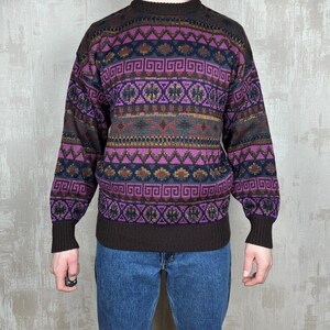 K&ouml;nnte beinhalten: Ein Vintage-Pullover mit geometrischem Muster in Lila, Braun und Gr&uuml;n. Der Pullover hat einen Rundhalsausschnitt und lange &Auml;rmel. Das Design zeigt sich wiederholende Formen und Linien.