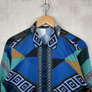 May include: A long-sleeved shirt with a geometric pattern in shades of blue, teal, black, and brown. The shirt is hanging on a wooden hanger. The collar and cuffs feature a repeating geometric design.