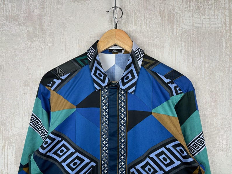 May include: A long-sleeved shirt with a geometric pattern in shades of blue, teal, black, and brown. The shirt is hanging on a wooden hanger. The collar and cuffs feature a repeating geometric design.