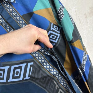 May include: A patterned button-up shirt in shades of blue, teal, black, and brown. The shirt features geometric and floral designs. The sleeve is visible, and a hand is partially visible, touching the shirt.