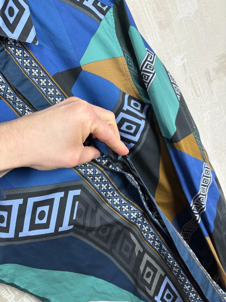 May include: A patterned button-up shirt in shades of blue, teal, black, and brown. The shirt features geometric and floral designs. The sleeve is visible, and a hand is partially visible, touching the shirt.