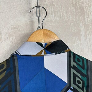 May include: A shirt with a geometric pattern in blue, white, black, and grey hangs on a wooden hanger. The collar of the shirt is visible, displaying the intricate design. The shirt is suspended from a metal chain.