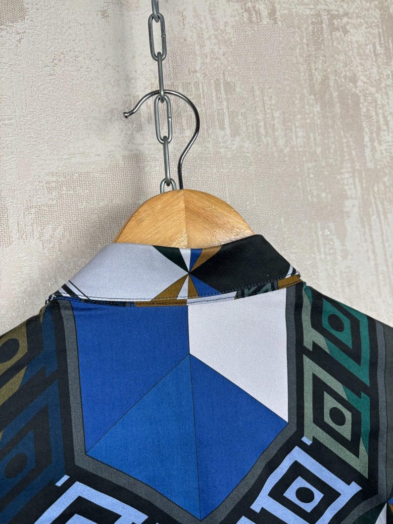 May include: A shirt with a geometric pattern in blue, white, black, and grey hangs on a wooden hanger. The collar of the shirt is visible, displaying the intricate design. The shirt is suspended from a metal chain.