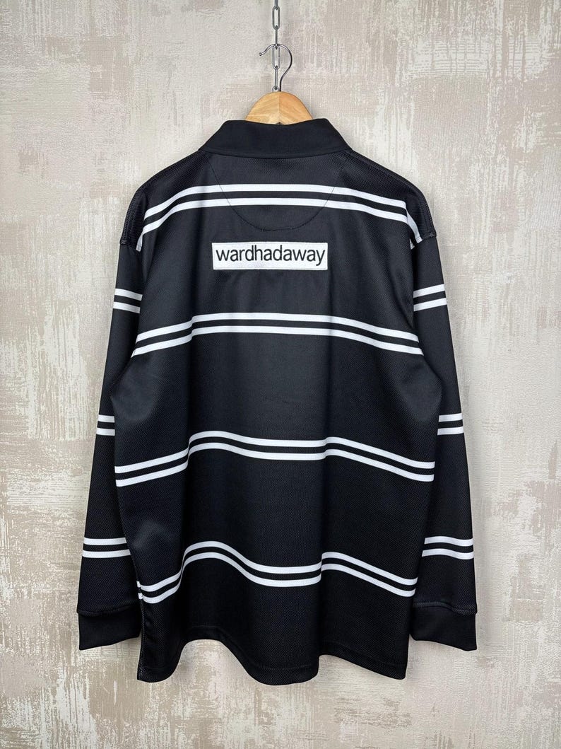 May include: Black long-sleeve shirt with white horizontal stripes. The shirt features a collared neckline and the text "wardhadaway" on the back. The shirt is hanging from a wooden hanger.