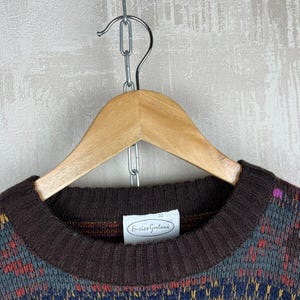 K&ouml;nnte beinhalten: Ein brauner Rundhals-Pullover mit einem farbenfrohen geometrischen Muster in Lila, Blau, Gelb und Rot. Der Pullover h&auml;ngt an einem Holzb&uuml;gel. Das Etikett lautet "Enrico Gorlani".