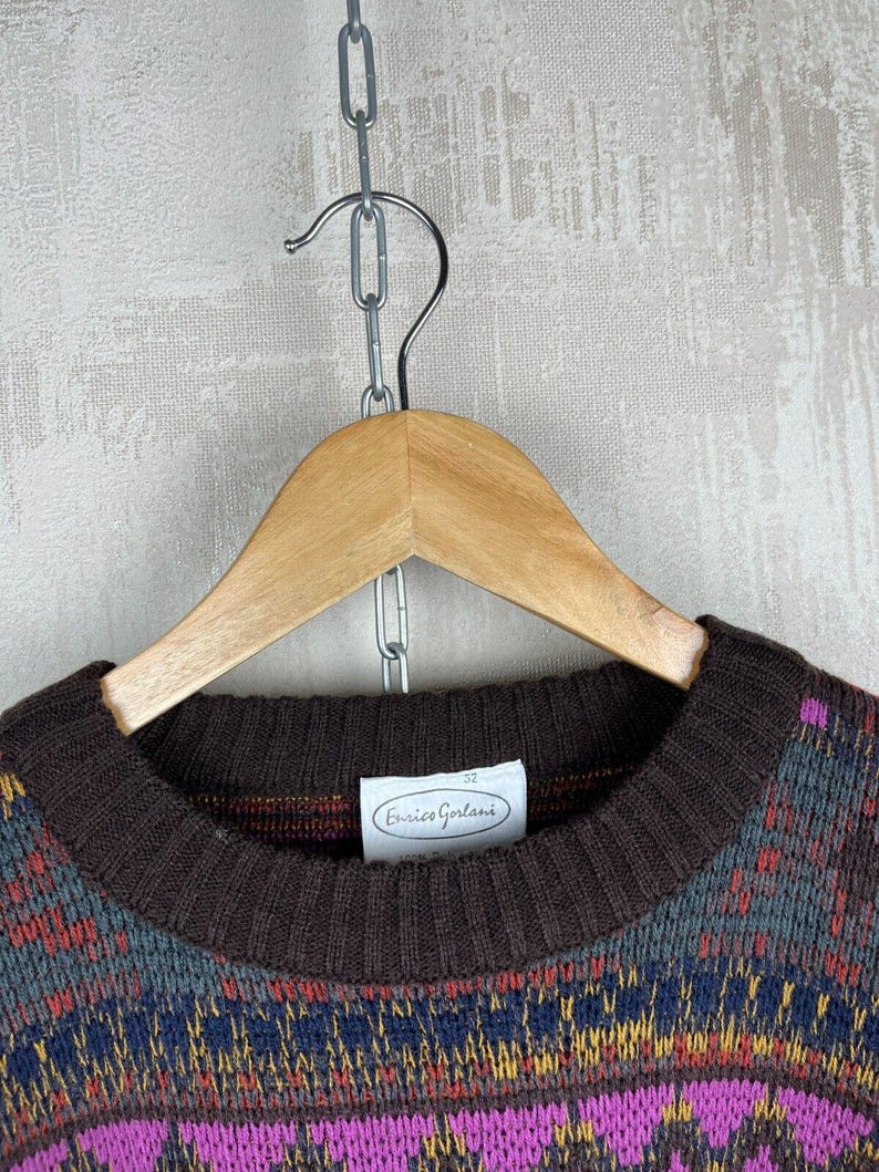 K&ouml;nnte beinhalten: Ein brauner Rundhals-Pullover mit einem farbenfrohen geometrischen Muster in Lila, Blau, Gelb und Rot. Der Pullover h&auml;ngt an einem Holzb&uuml;gel. Das Etikett lautet "Enrico Gorlani".