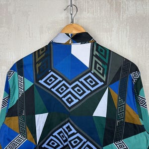 May include: A patterned button-down shirt in blue, teal, brown, and black. The geometric design features diamond and rectangular shapes. The shirt is hanging from a wooden hanger.