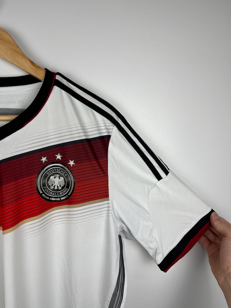 May include: White soccer jersey with black and red stripes and the German national team logo, "Deutscher Fussball-Bund", with an eagle inside a circle.